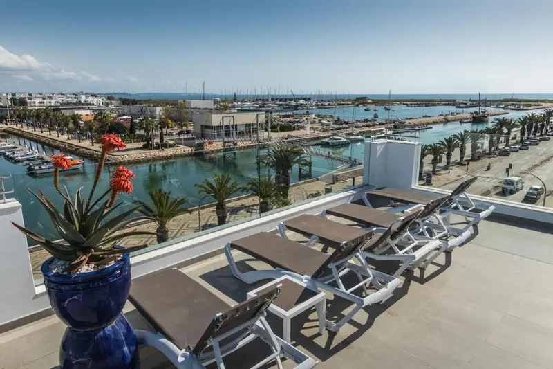 Transats blancs alignés sur une terrasse ensoleillée avec vue sur un port de plaisance.