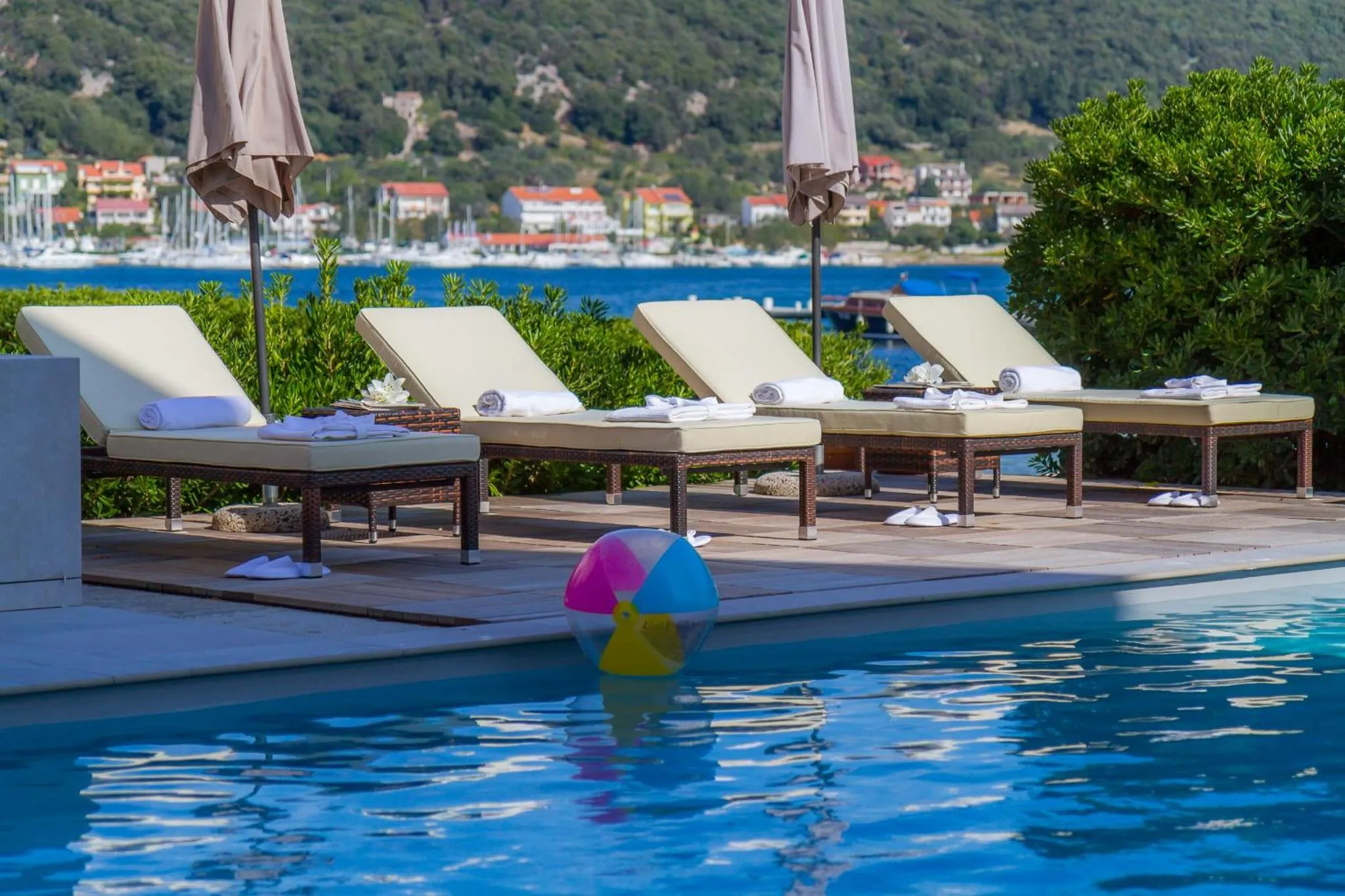 Transats blancs alignés au bord d'une piscine turquoise, avec une vue sur la mer et des collines verdoyantes.