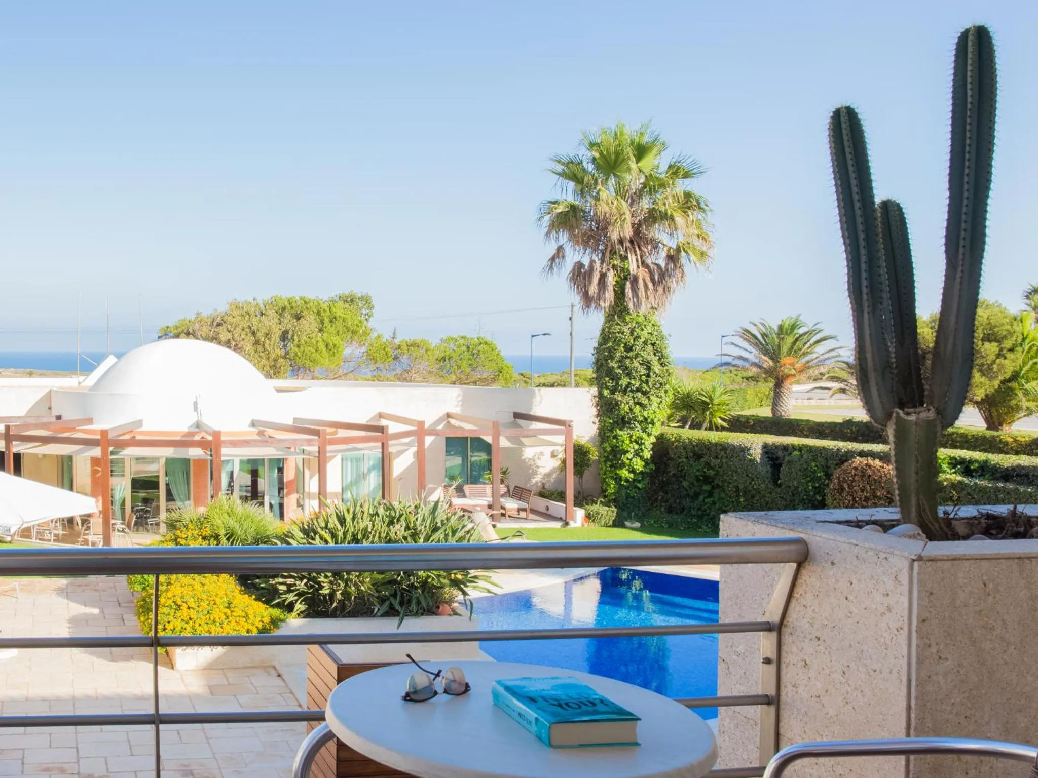 Une terrasse avec une table ronde et une piscine au premier plan, avec une végétation luxuriante et une vue sur la mer.