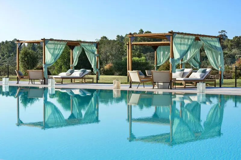 Transats et pergolas ombragées bordent une piscine turquoise reflétant le ciel bleu.