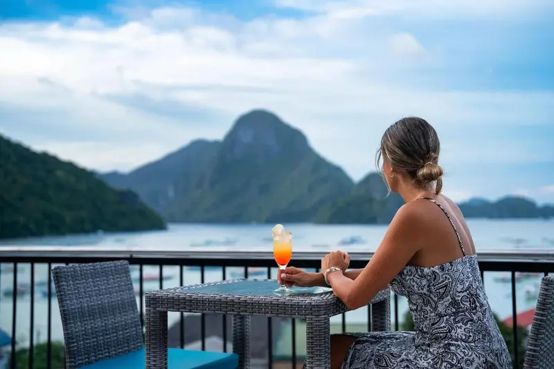 Une personne est assise à une table avec des boissons sur une terrasse avec vue sur la mer et les montagnes.