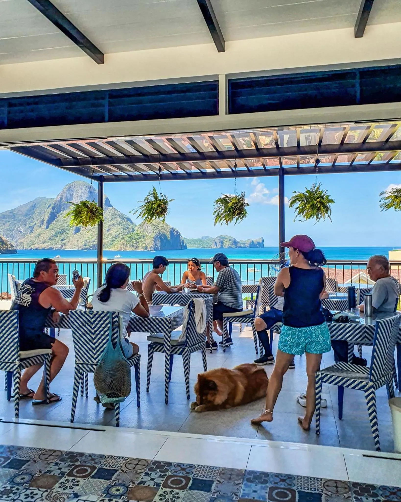 Des convives sont assis à des tables sur une terrasse ombragée avec vue sur la mer.