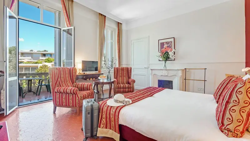 Une chambre lumineuse avec un lit et deux fauteuils près d'une fenêtre ouverte sur un balcon.