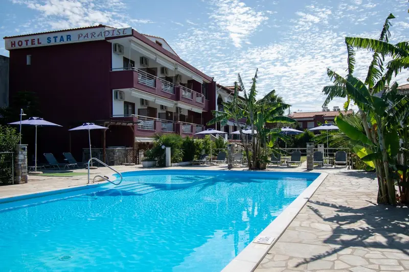 Piscine rectangulaire devant un bâtiment à plusieurs étages avec des balcons et des parasols.