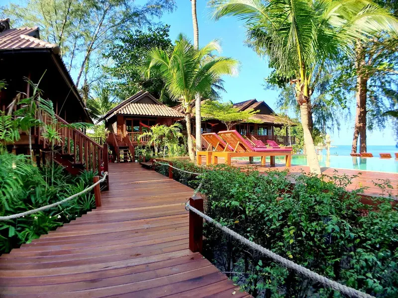 Allée en bois menant à des bungalows sur pilotis au milieu d'une végétation tropicale.