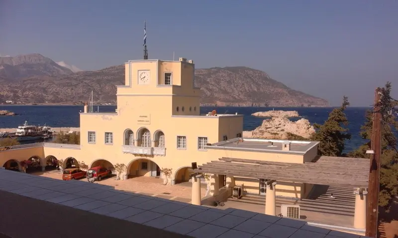 Bâtiment jaune à colonnes avec une horloge, surplombant un port de plaisance et des montagnes.