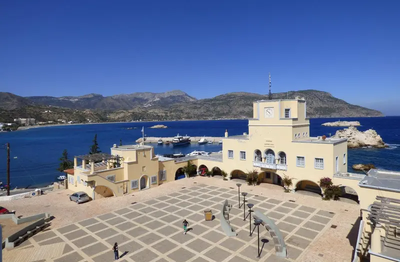 Vue d'un bâtiment beige et blanc avec une place pavée en contrebas et la mer en arrière-plan.