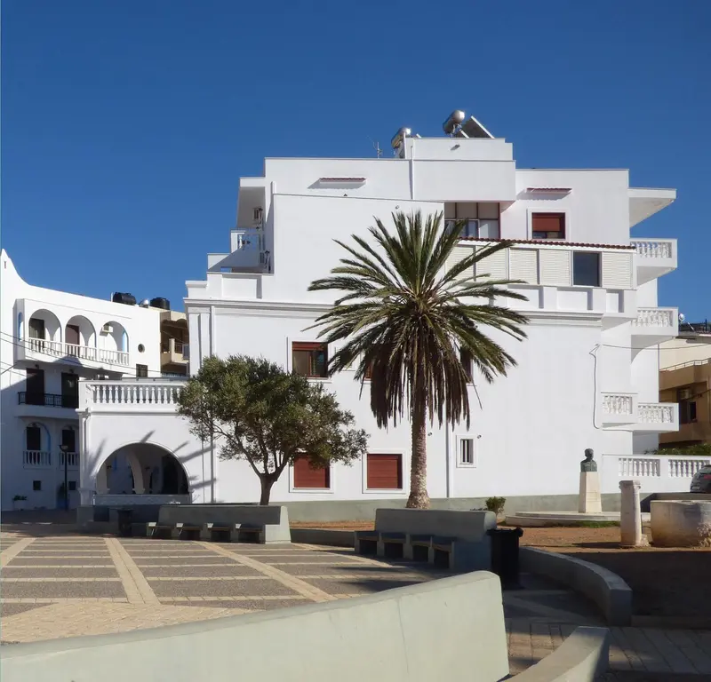 Bâtiment blanc à plusieurs étages avec des balcons et un palmier devant.