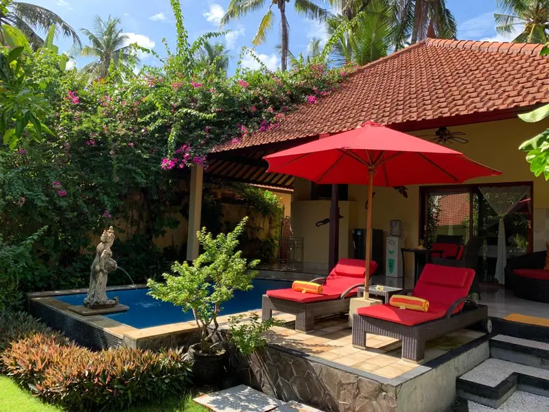 Petite piscine et transats sous un parasol rouge devant un bâtiment jaune.