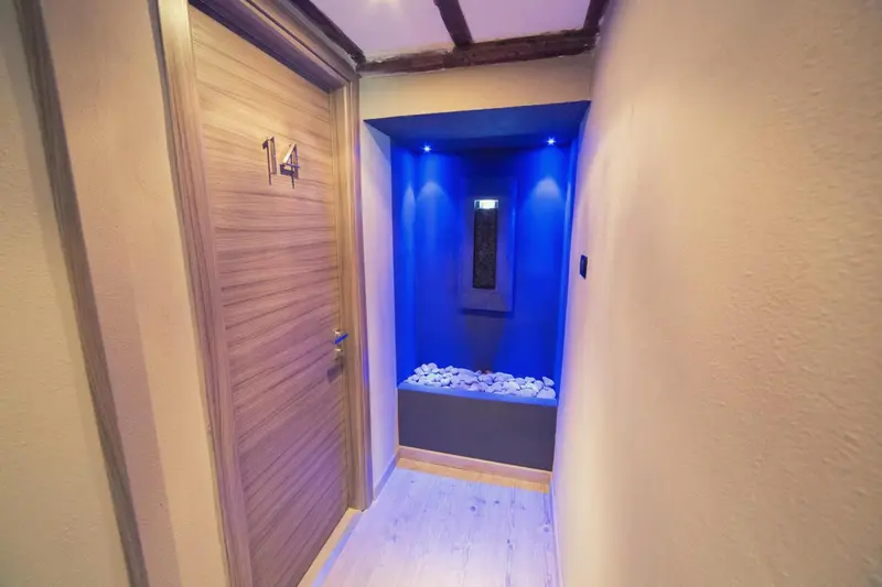 Un couloir d'hôtel mène à une porte numérotée et à un espace éclairé de bleu.