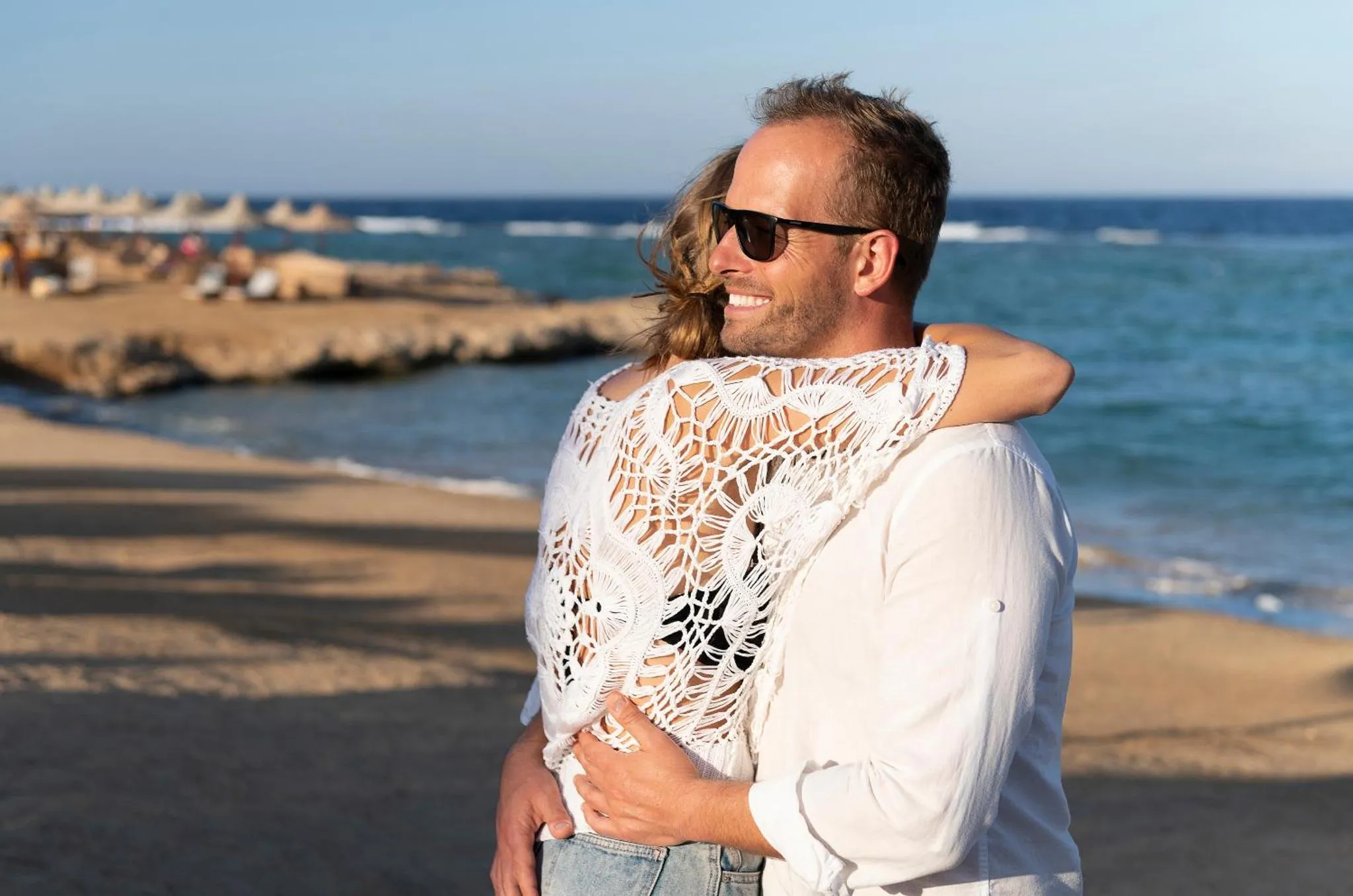 Un couple souriant se tient sur une plage de sable fin, face à la mer.