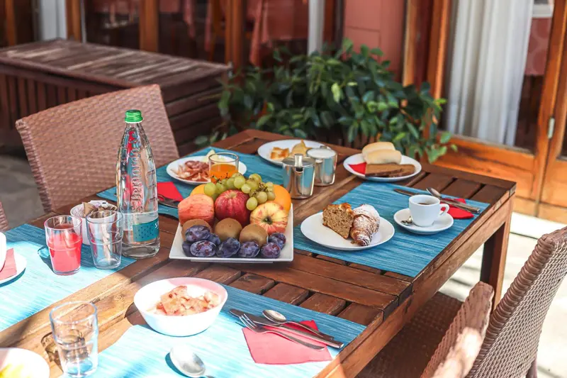Une table en bois est dressée avec un petit-déjeuner composé de fruits, de viennoiseries et de boissons.