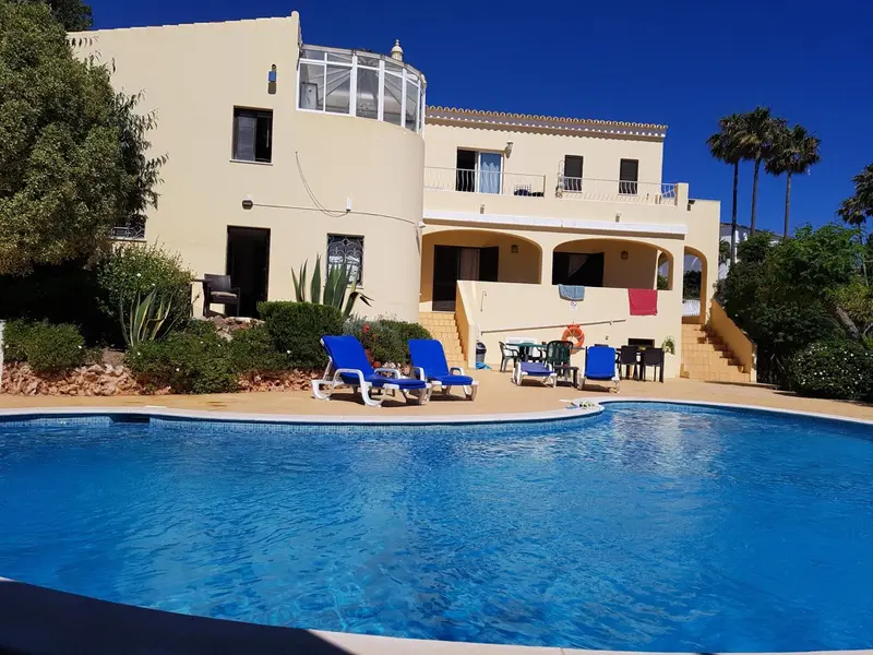Une piscine turquoise au premier plan, avec des transats bleus et un bâtiment jaune à l'arrière-plan.