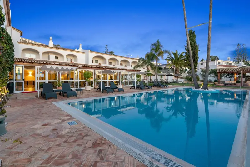 Piscine turquoise bordée d'une terrasse ensoleillée devant un bâtiment blanc.