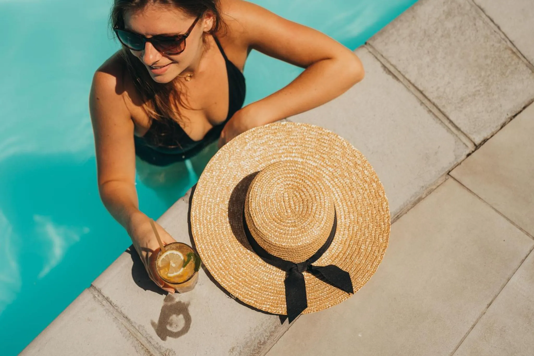 Une femme se détend au bord d'une piscine avec un chapeau et une boisson.