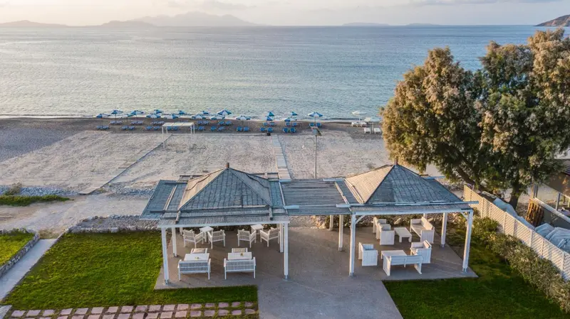 Terrasse ombragée avec tables et chaises donnant sur une plage de sable et la mer.