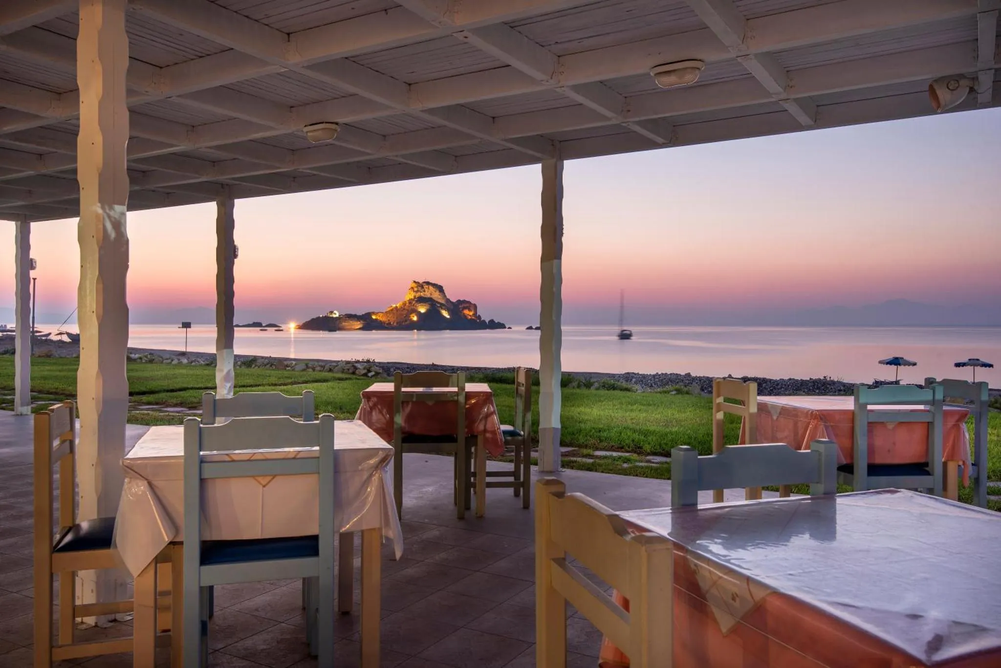 Terrasse ombragée avec tables et chaises donnant sur la mer au coucher du soleil.