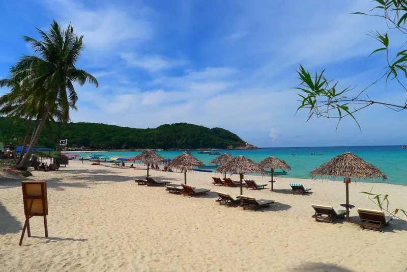 Plage de sable blanc bordée de parasols et de chaises longues, avec une mer turquoise en arrière-plan.