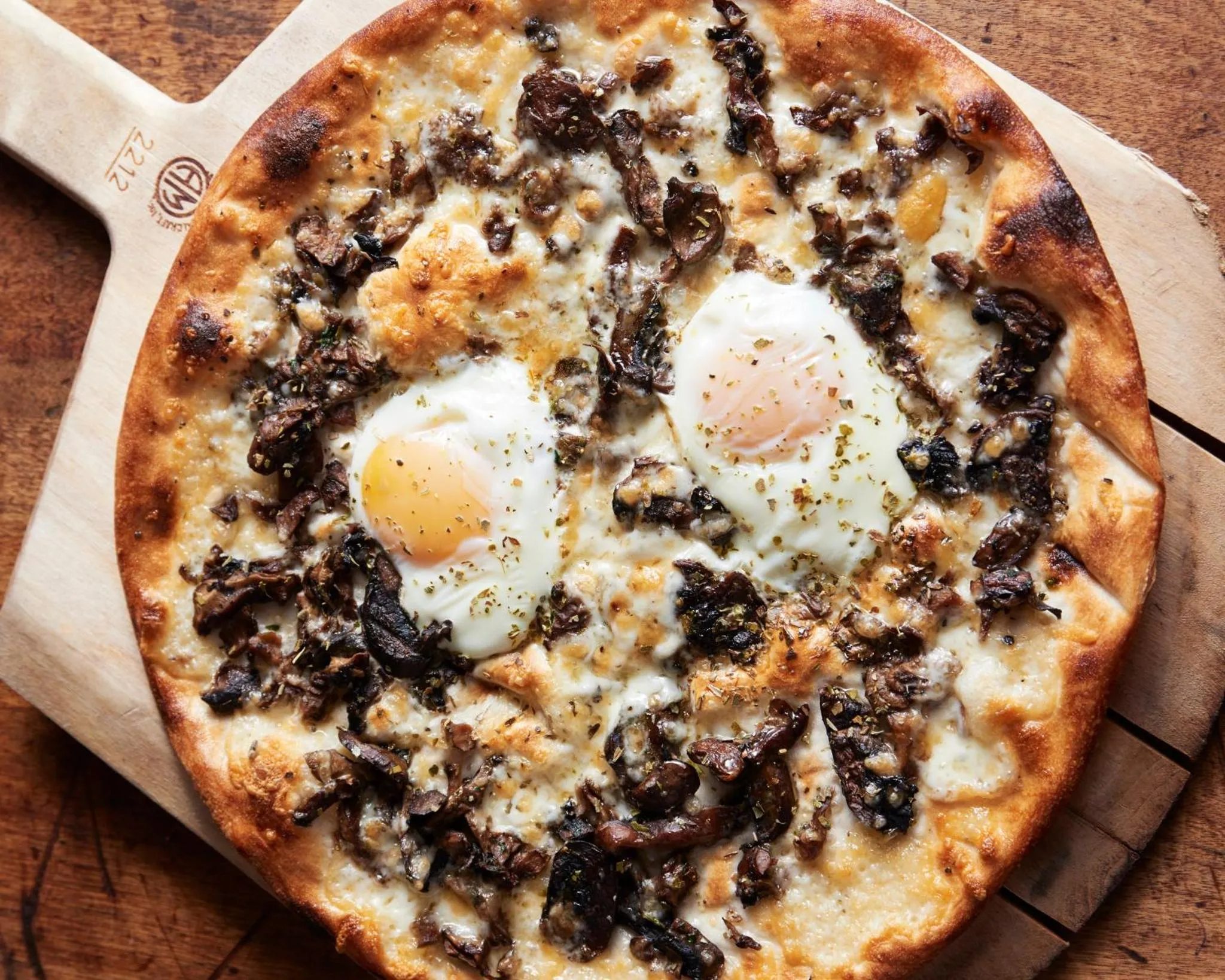 Une pizza aux champignons et aux œufs au plat est présentée sur une planche à pizza en bois.