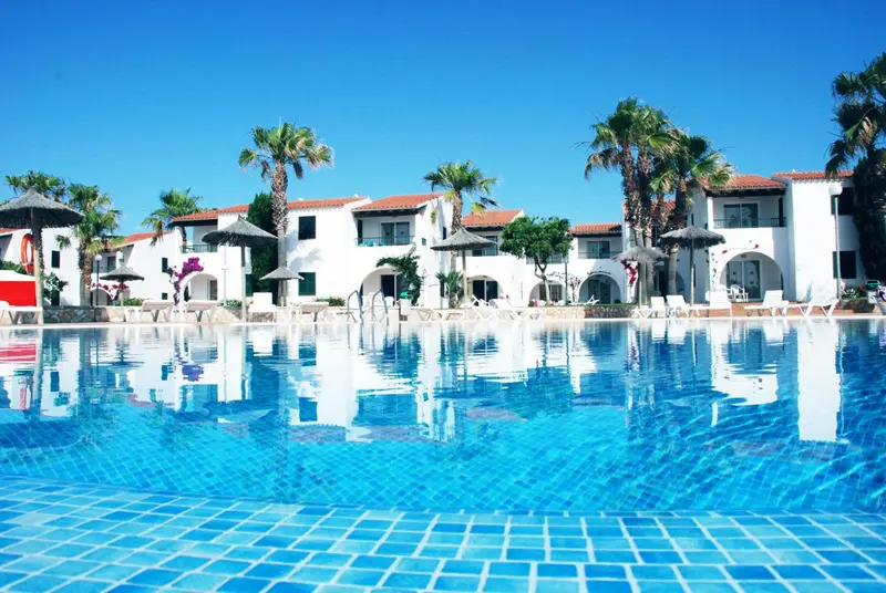 Vue d'une piscine turquoise reflétant un bâtiment blanc et des palmiers sous un ciel bleu.