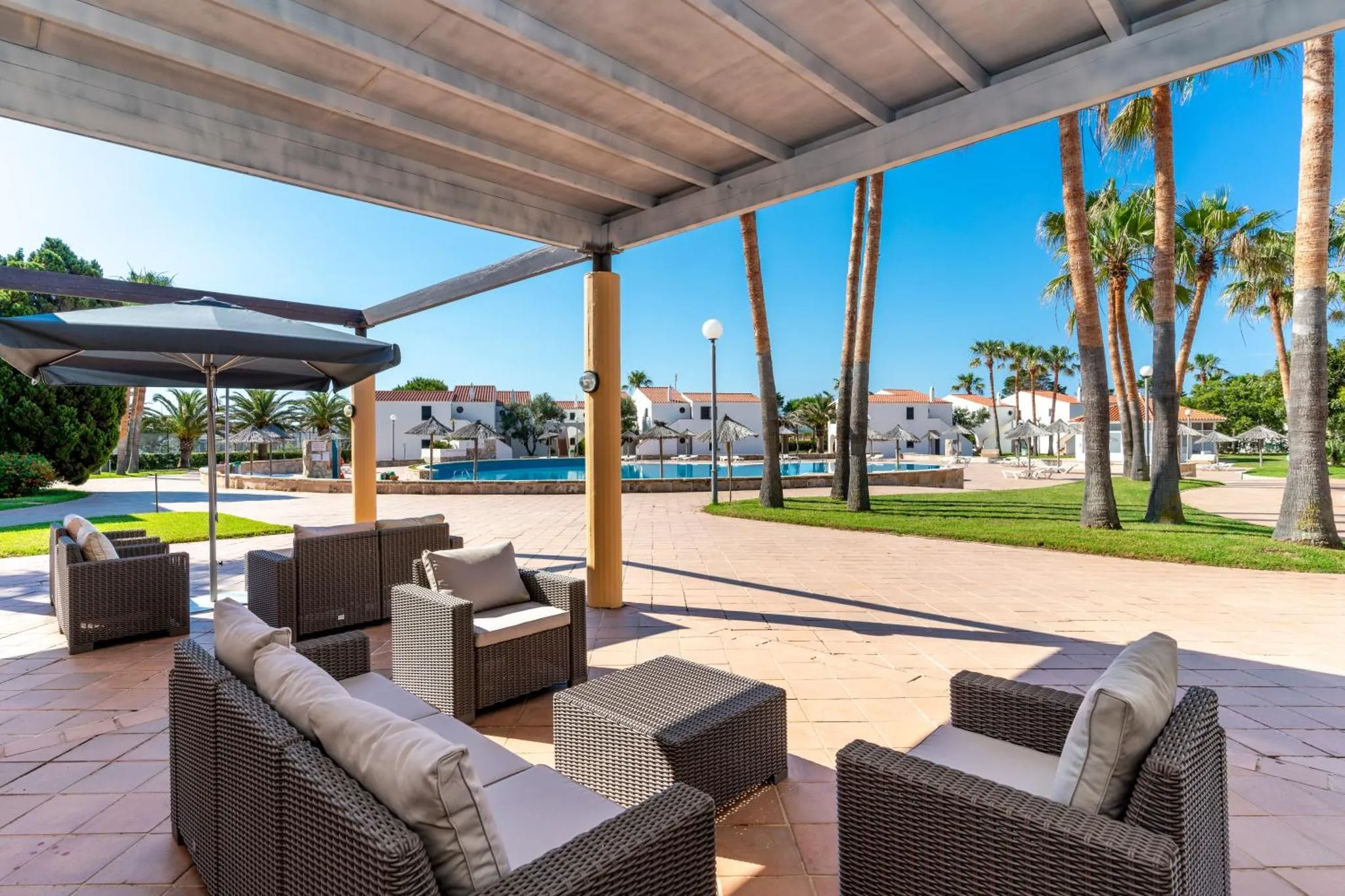 Espace de détente extérieur avec mobilier en résine tressée et vue sur une piscine et des palmiers.