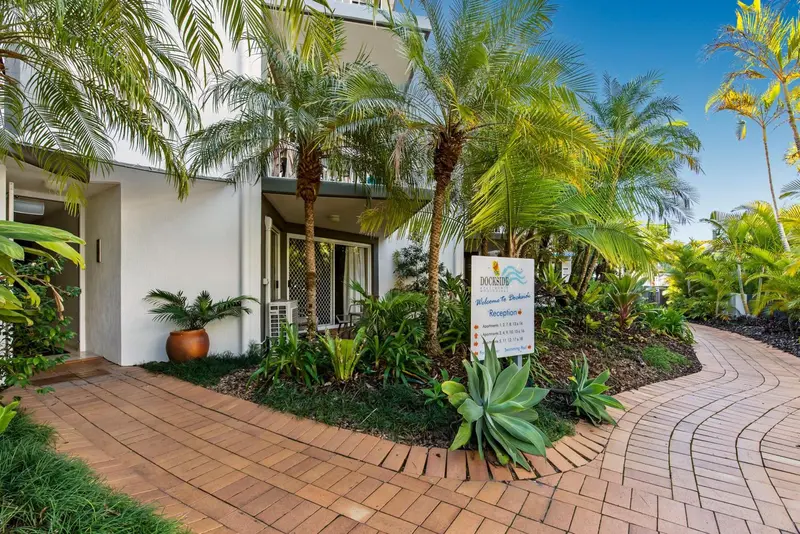 Entree d'un batiment blanc avec un jardin tropical et un panneau d'accueil.