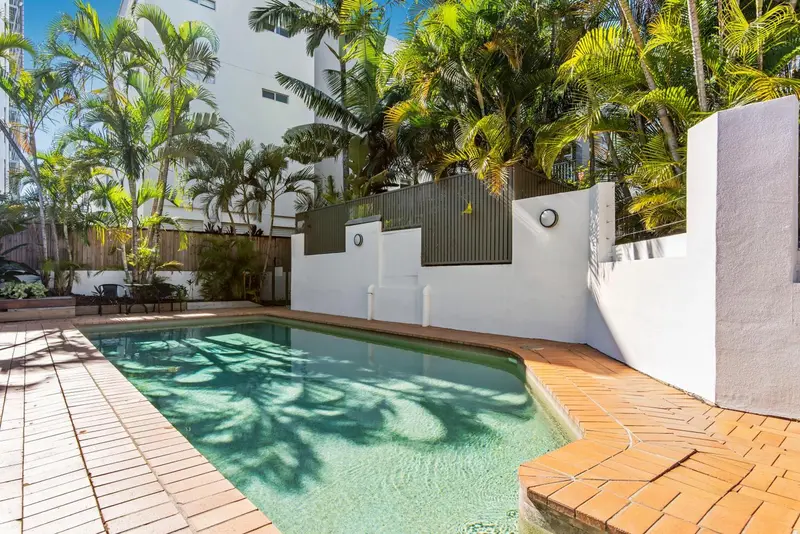 Une piscine rectangulaire bordée d'une terrasse carrelée, entourée de végétation tropicale et d'un mur blanc.