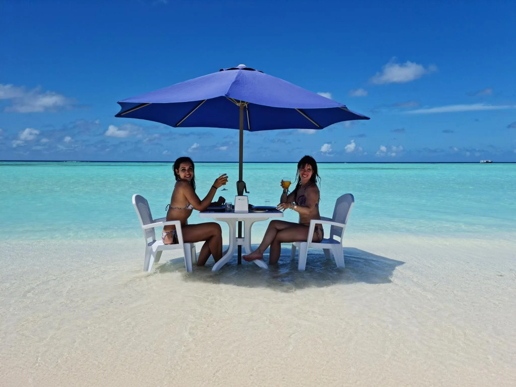 Deux femmes sourient en sirotant des boissons à une table sur une plage de sable blanc.