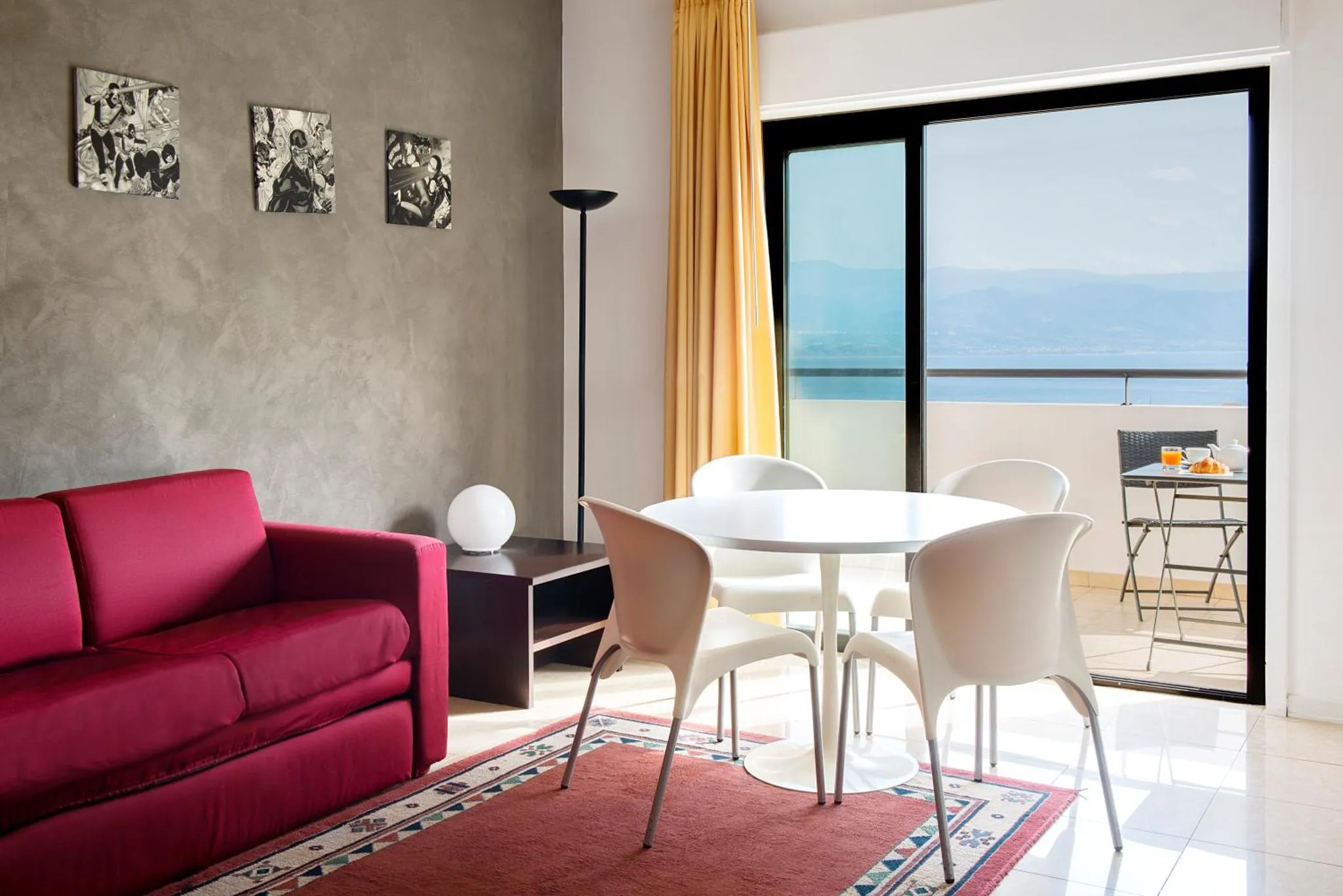 Espace de vie intérieur avec canapé rouge, table et chaises près d'une baie vitrée donnant sur la mer.