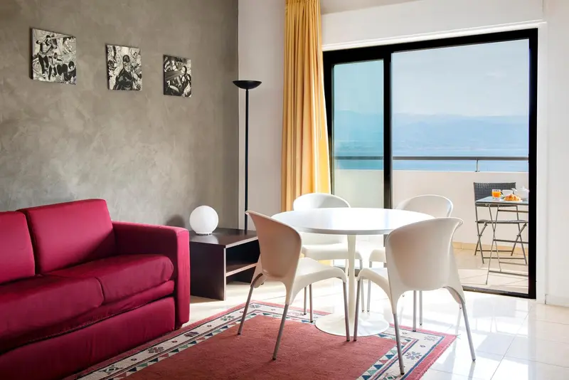Espace de vie intérieur avec canapé rouge, table et chaises près d'une baie vitrée donnant sur la mer.