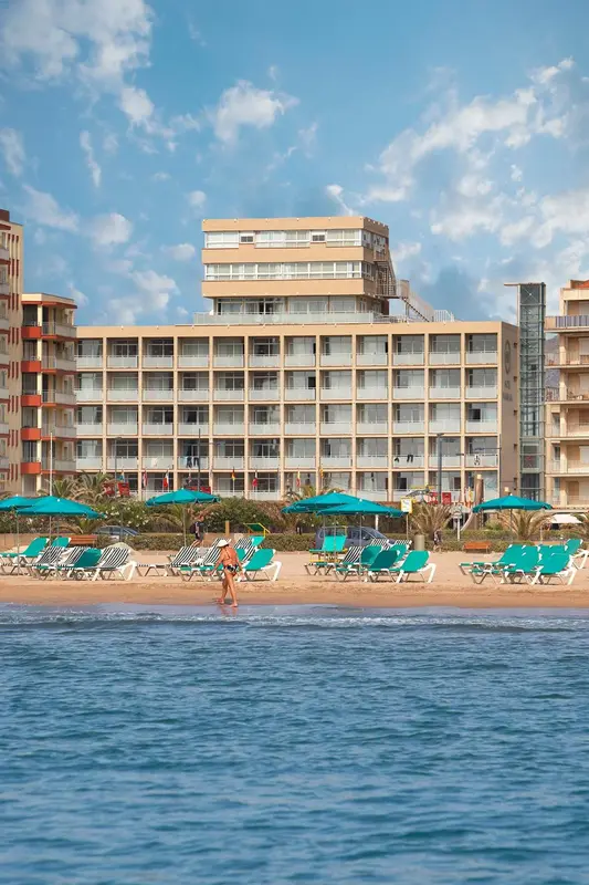 Vue d'une plage de sable fin avec des transats bleus devant un bâtiment hôtelier.