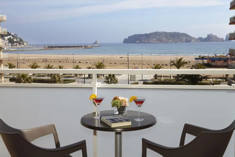 Une table ronde avec des boissons et des fleurs sur un balcon avec vue sur la mer et la plage.