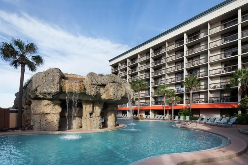 Une piscine turquoise avec une cascade rocheuse et des transats est devant un bâtiment à plusieurs étages.