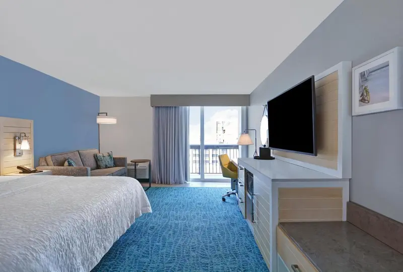 Chambre lumineuse avec un grand lit et un bureau près d'une baie vitrée.