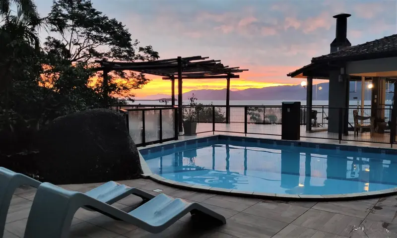Une piscine avec transats surplombe une mer calme au coucher du soleil.