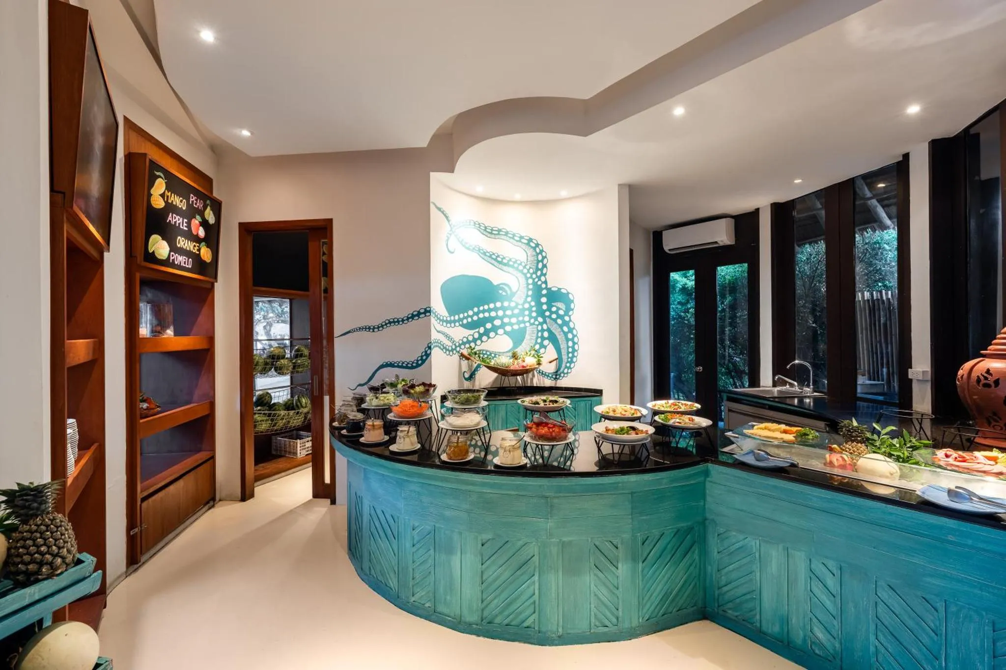 Un buffet de fruits et de viennoiseries est présenté sur un comptoir turquoise dans une salle lumineuse.