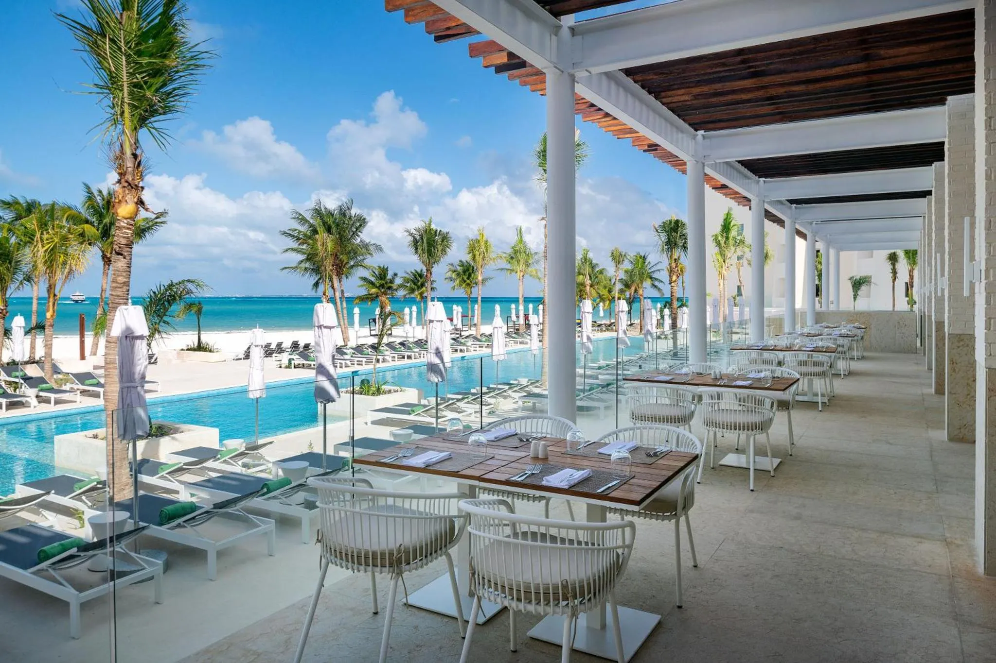 Terrasse ombragée donnant sur une piscine et une plage de sable blanc.