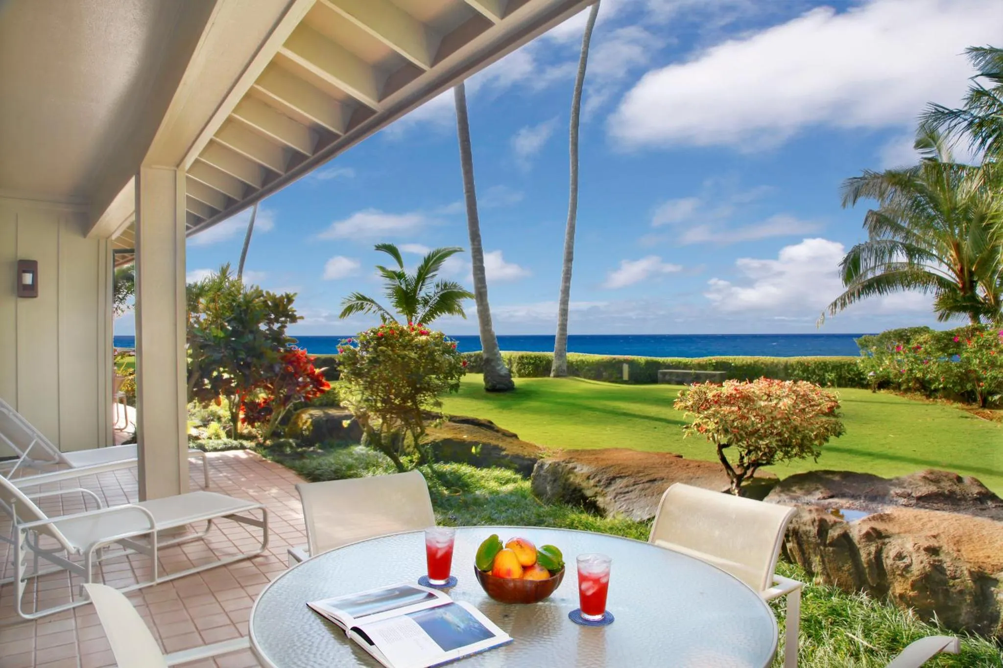 Une table ronde avec des fruits et des verres est placée sur une terrasse ensoleillée avec vue sur la mer.