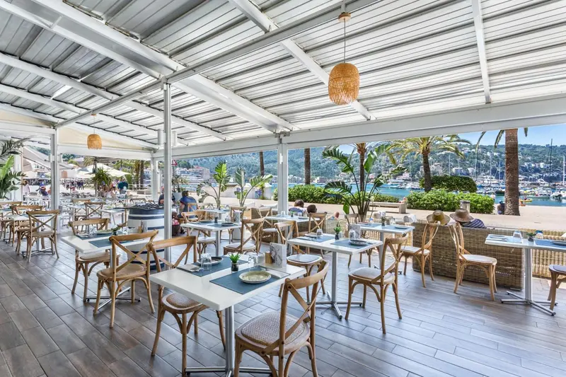 Terrasse ombragée avec tables et chaises donnant sur un port de plaisance.