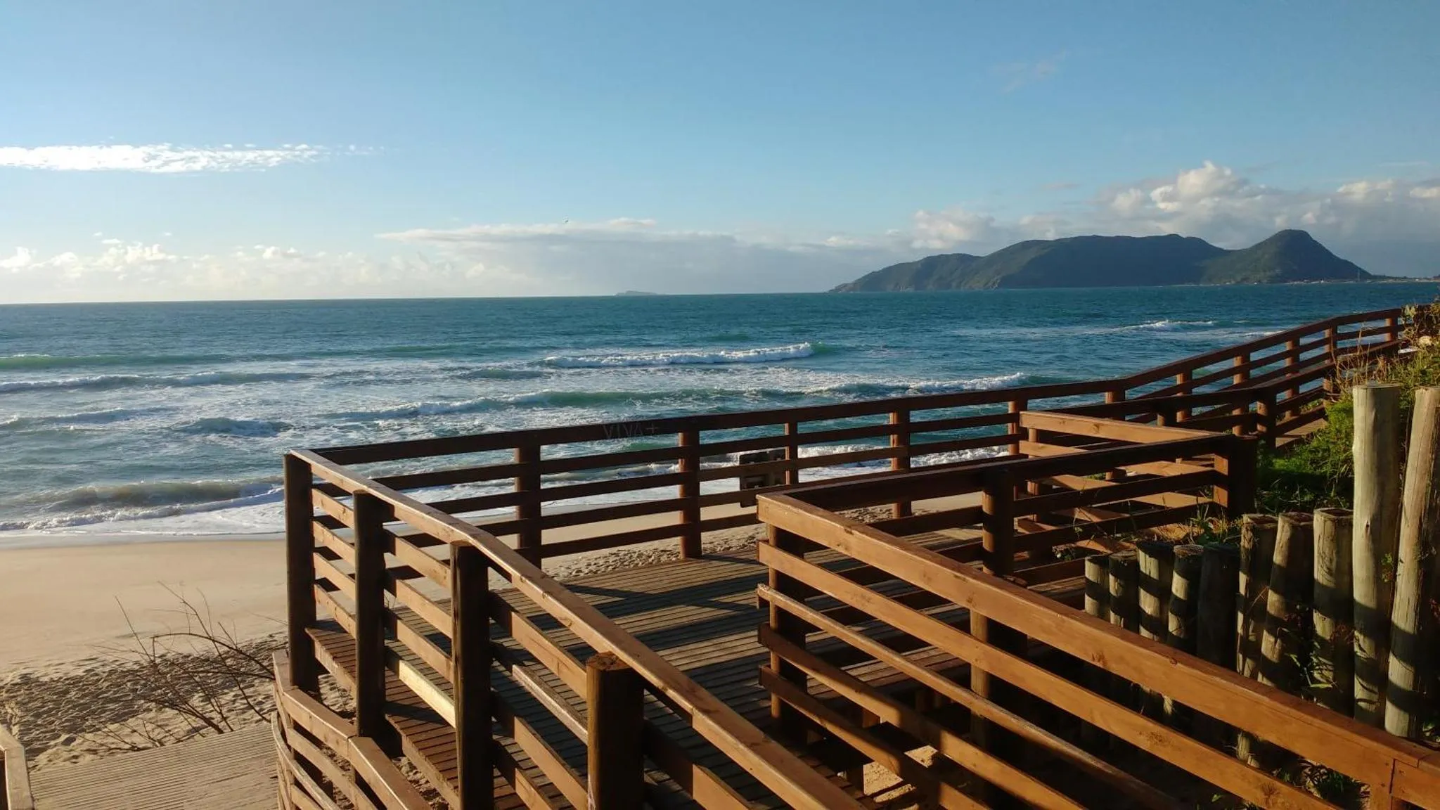 Une promenade en bois descend vers une plage de sable fin avec une vue sur la mer et des collines au loin.