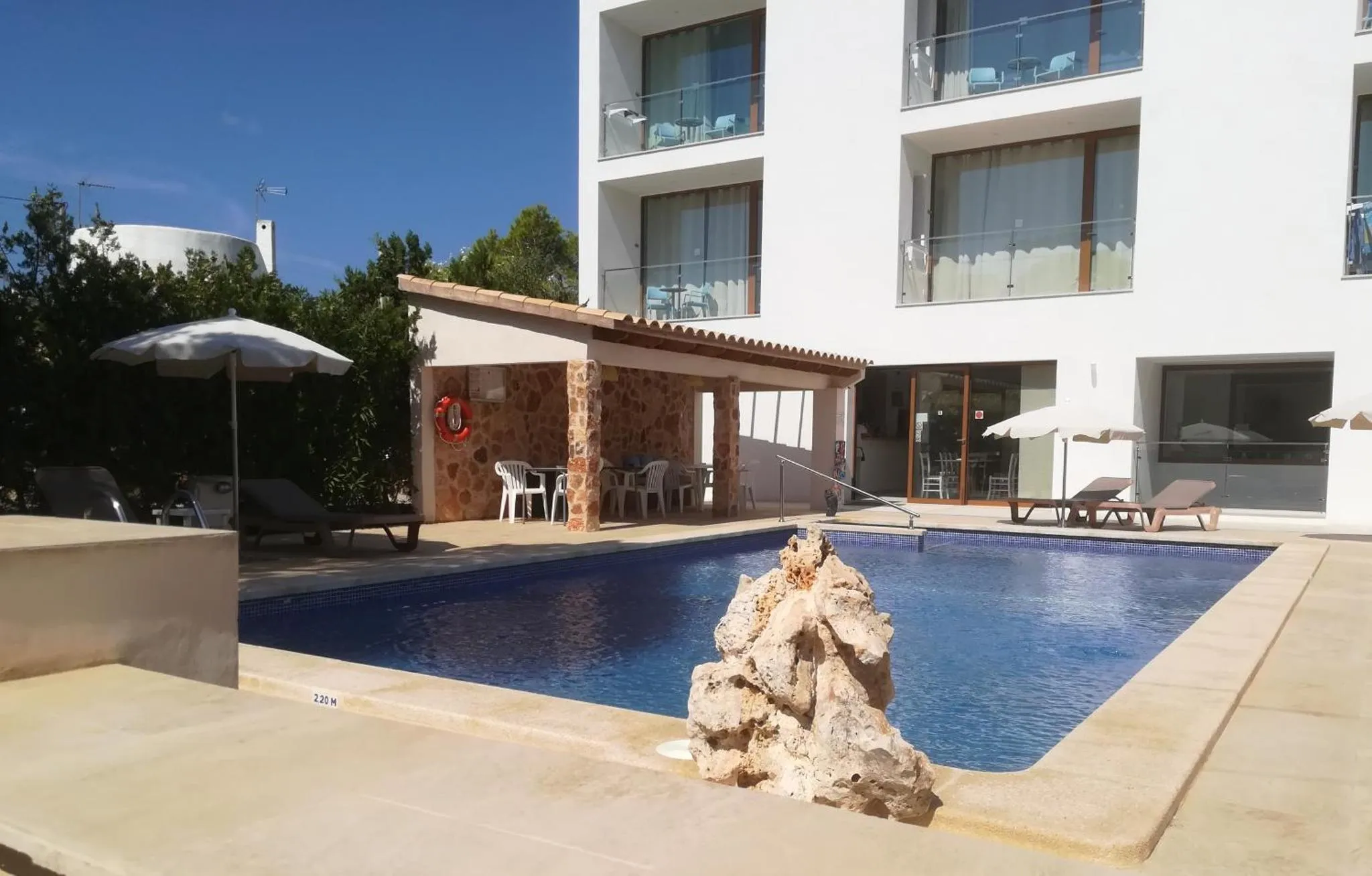Une piscine rectangulaire avec un rocher au centre, entourée d'une terrasse avec des transats et des parasols.