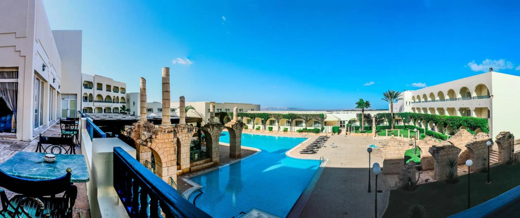 Vue d'une piscine d'hôtel avec des colonnes et des palmiers sous un ciel bleu.