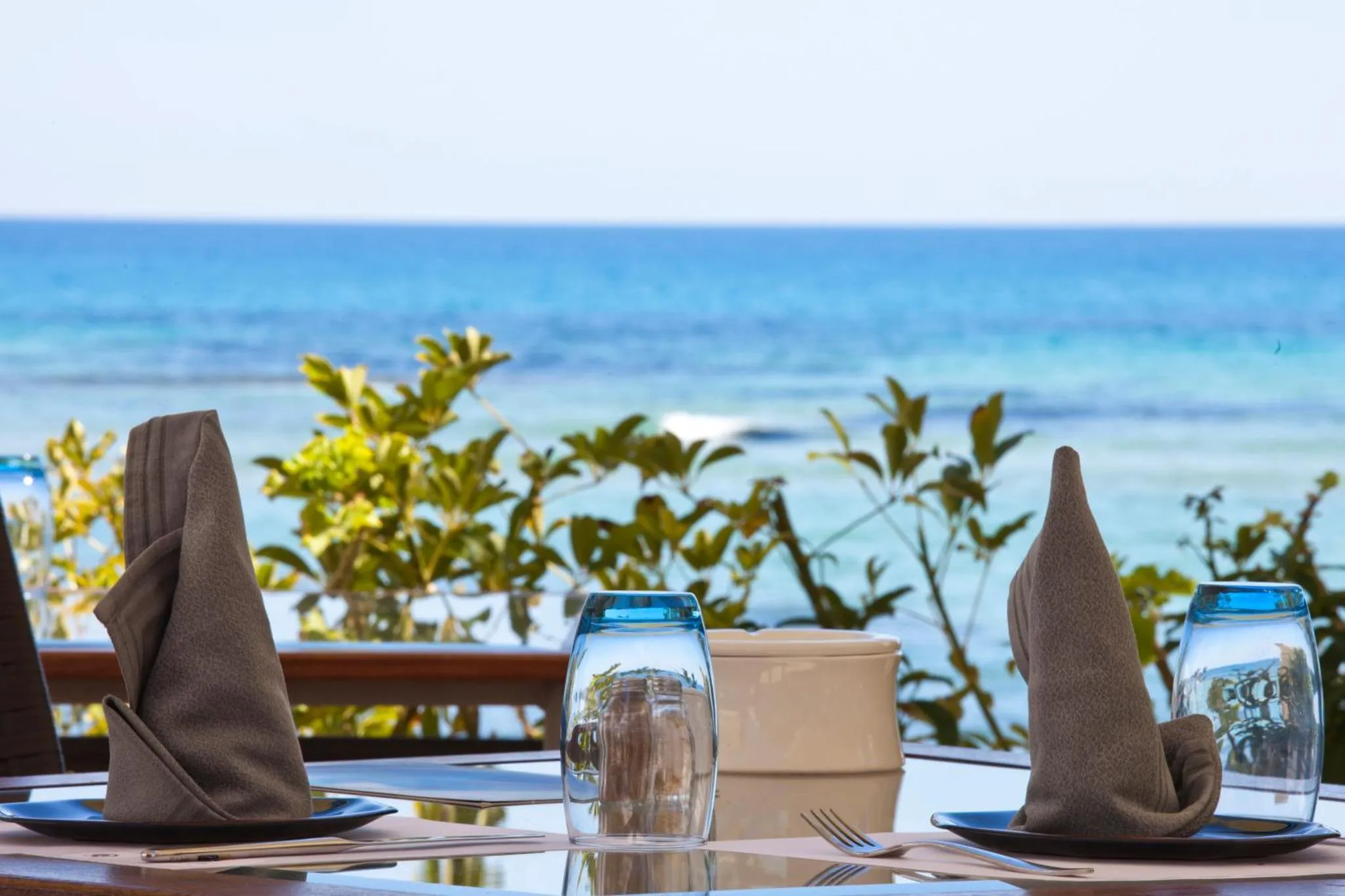 Une table est dressée avec des couverts et des verres devant une vue sur la mer.