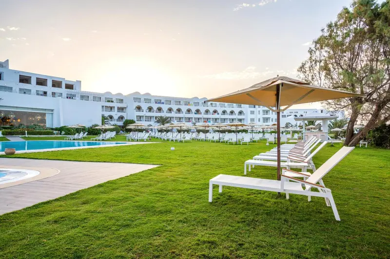 Transats blancs alignés sur une pelouse verte devant un bâtiment blanc et une piscine.