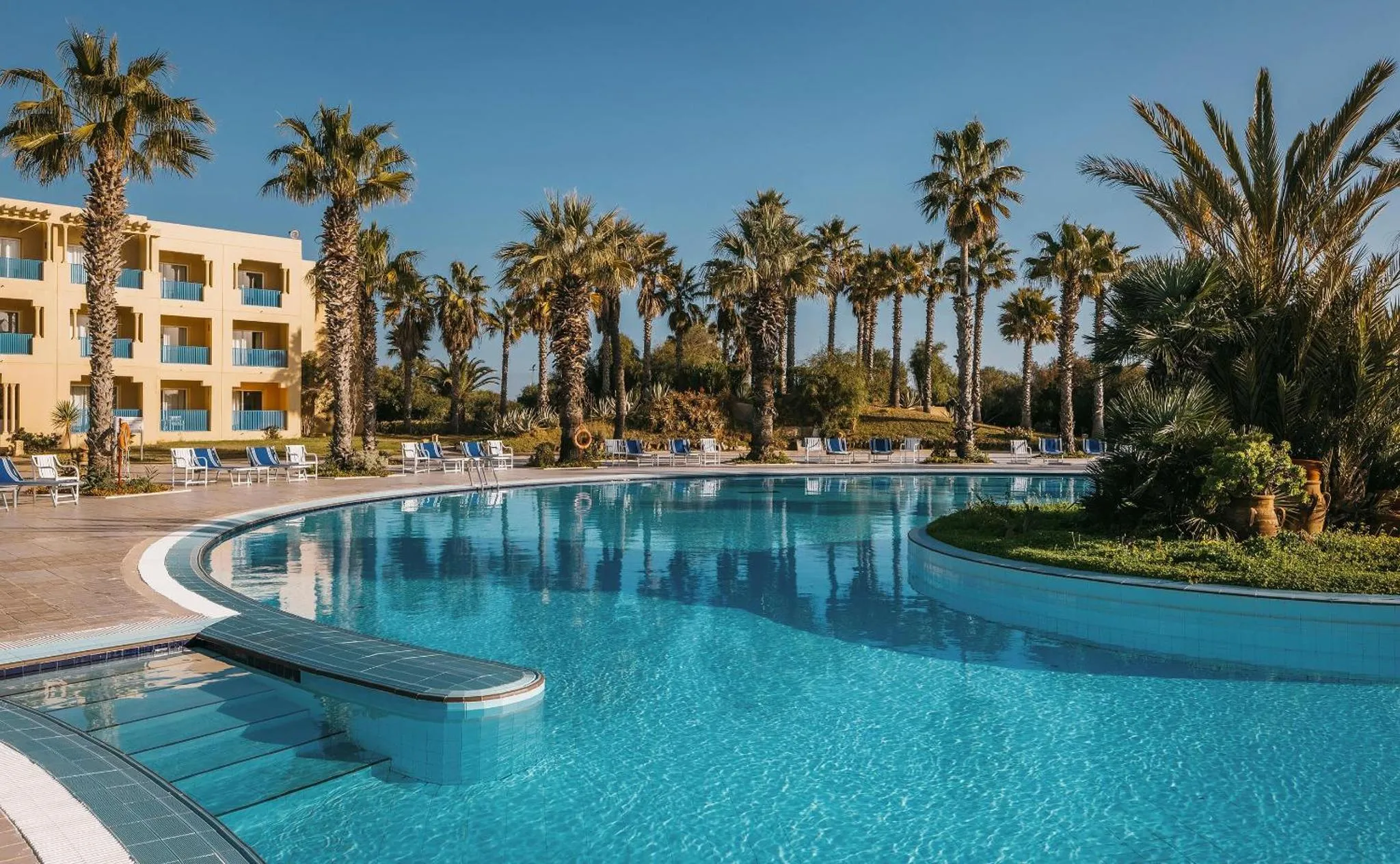 Piscine d'hôtel avec palmiers et transats sous un ciel bleu.