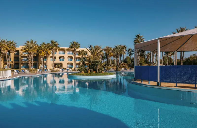 Une piscine turquoise reflète un bâtiment beige et des palmiers sous un ciel bleu.