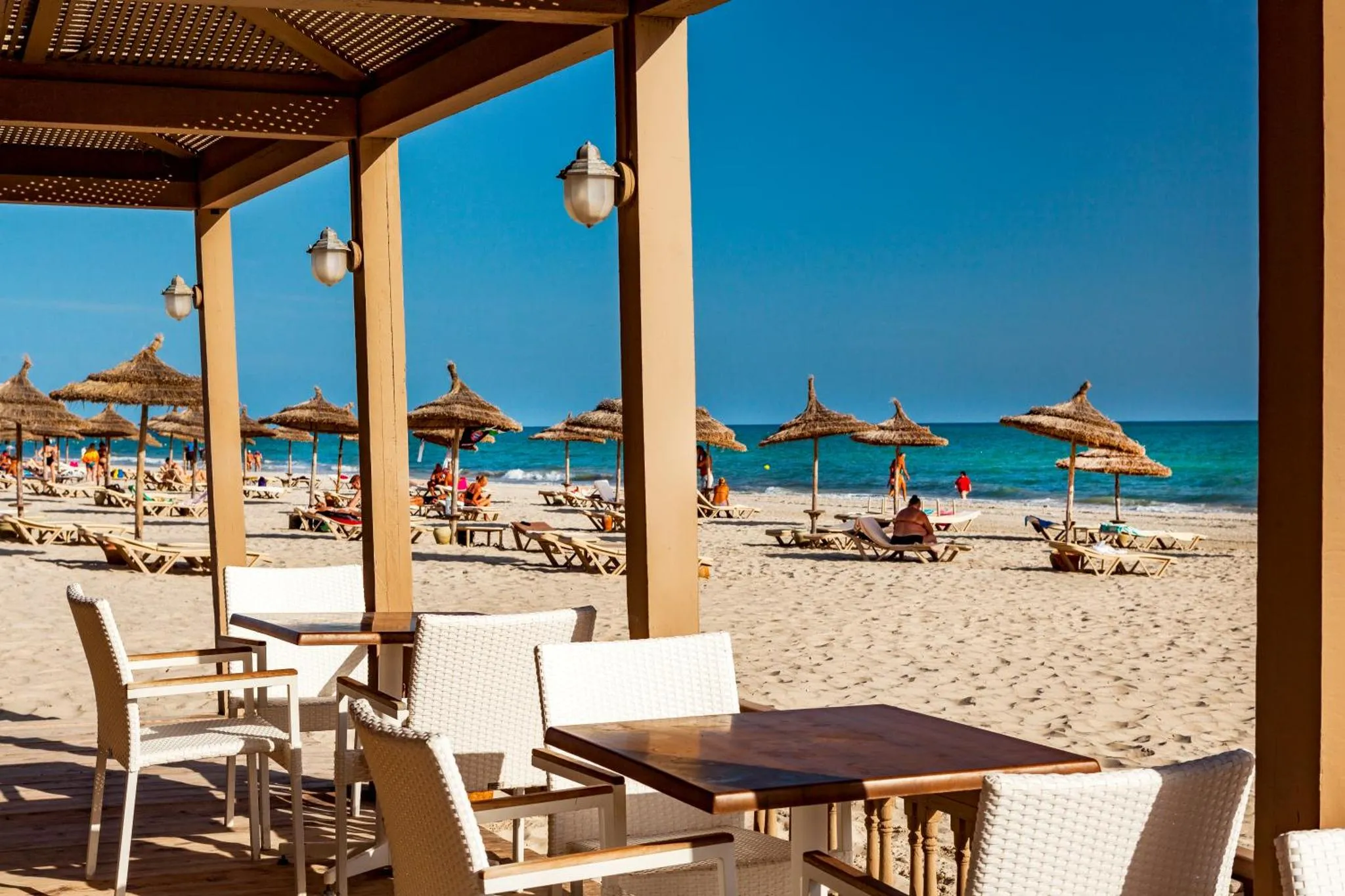 Terrasse ombragée donnant sur une plage ensoleillée avec parasols et transats.