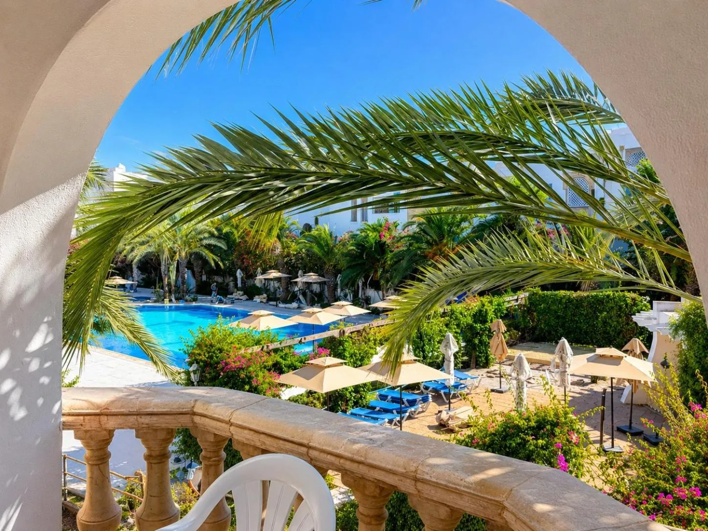 Vue sur une piscine ensoleillée depuis un balcon ombragé par des palmiers.
