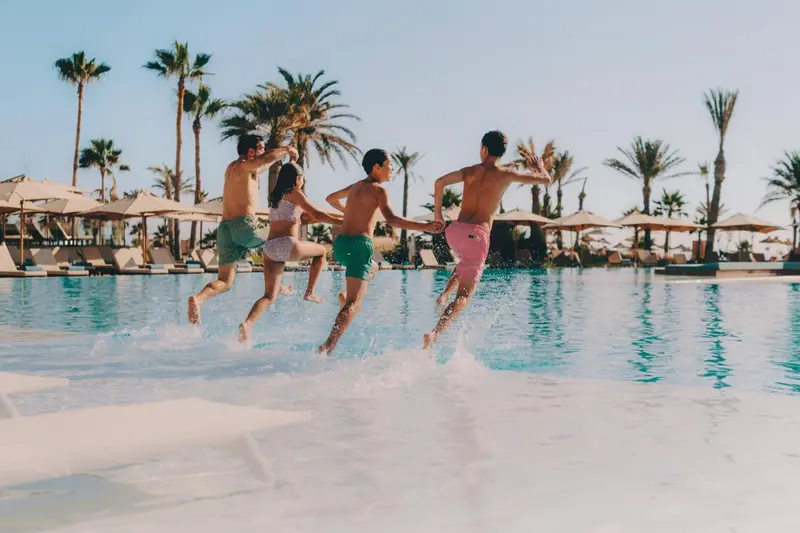 Des personnes sautent dans une piscine ensoleillée bordée de palmiers.