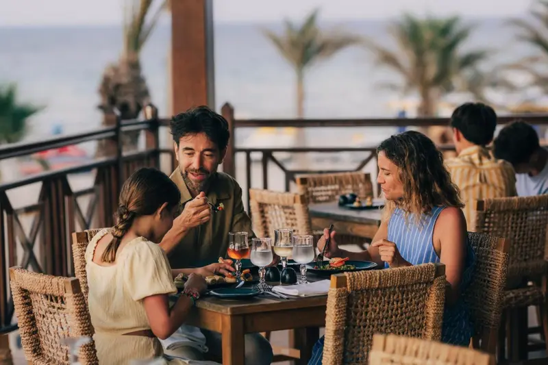 Une famille prend son repas sur une terrasse ensoleillée avec vue sur la mer.